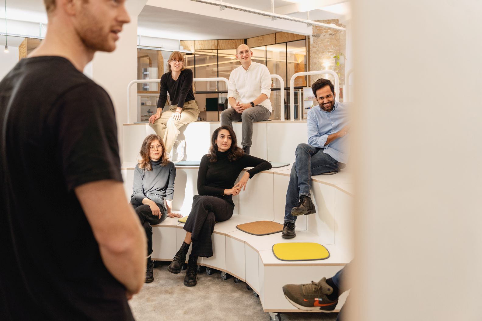 What if We Fly Taskboard: Gruppe von Mitarbeitern sitzt auf einer modularen, weißen Medienwand von Interstuhl.