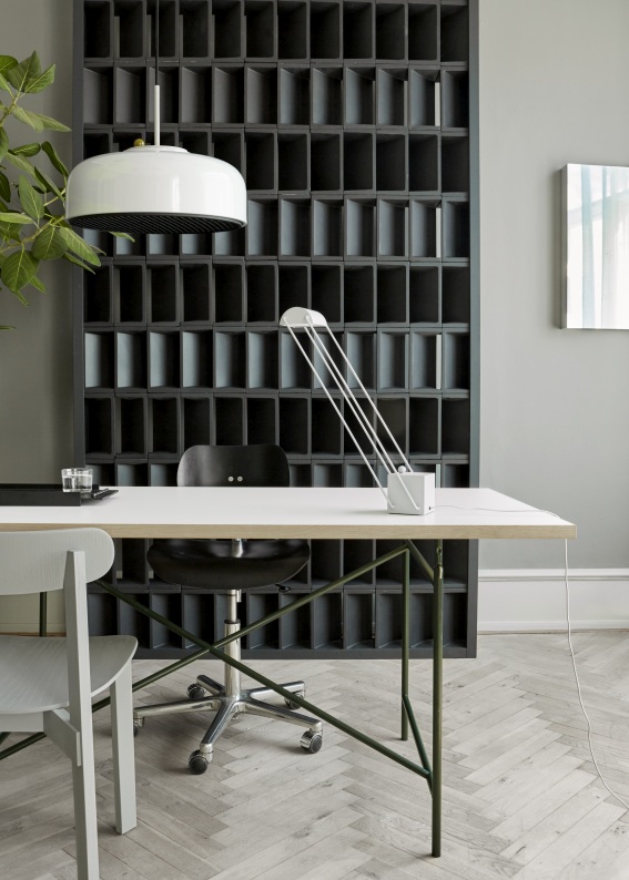 Moderner Schreibtisch mit weißer Tischplatte, grünen Beinen und weißer Lampe vor einem dunklen Regal.