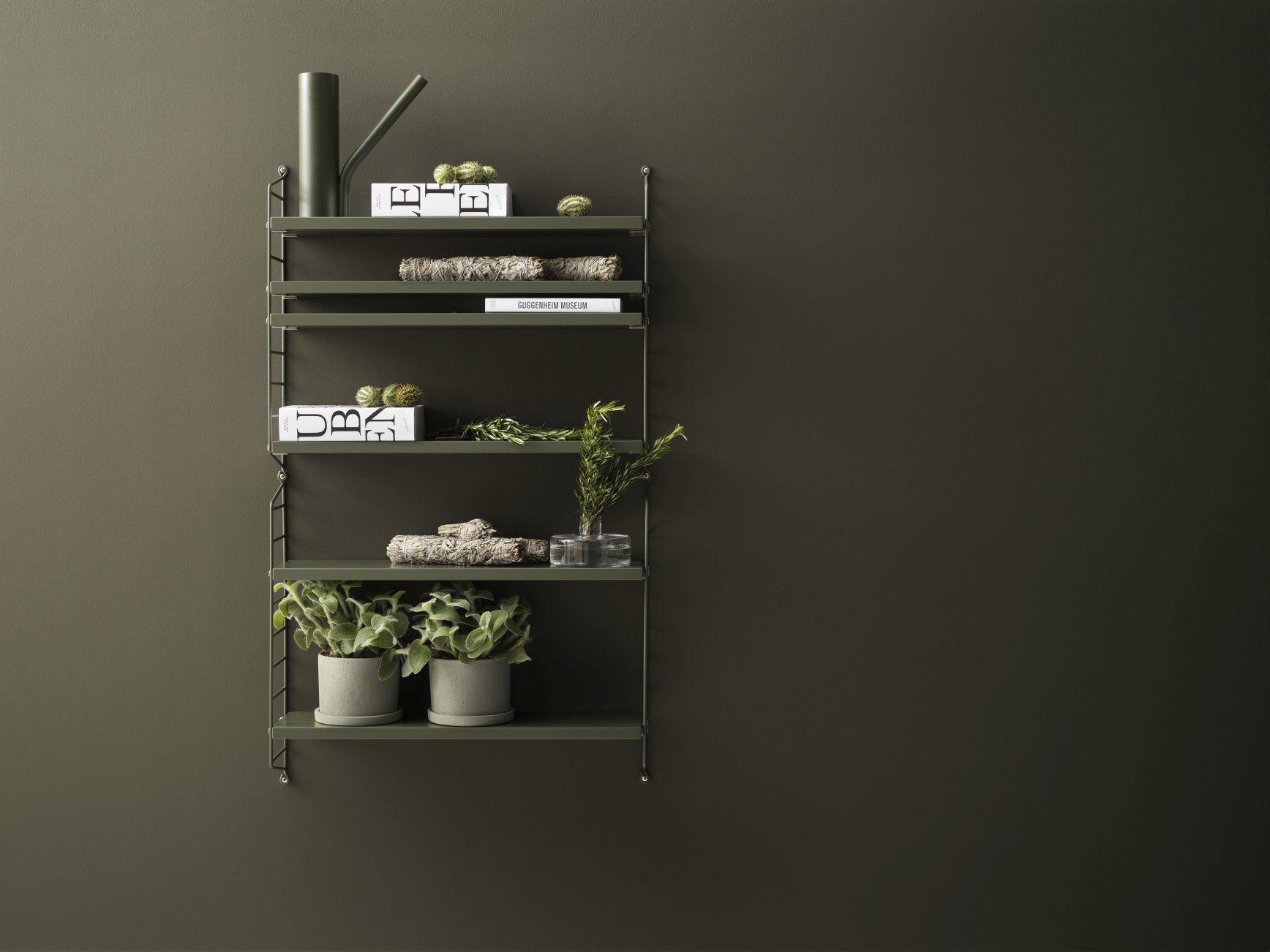 Grünes String Regal mit Dekoration an einer dunklen Wand. String Pocket Regal von String Furniture.