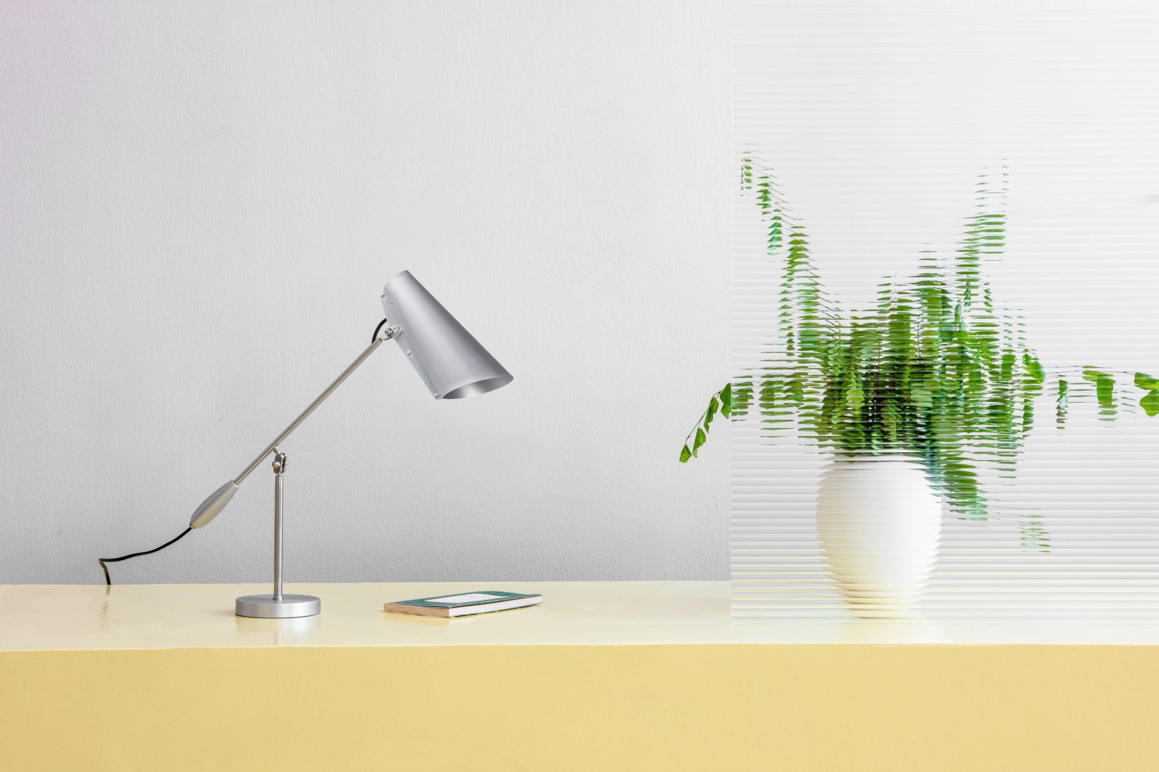 Silberne Birdy Tischleuchte auf gelbem Tisch mit Pflanze im Hintergrund. Design Lampe.