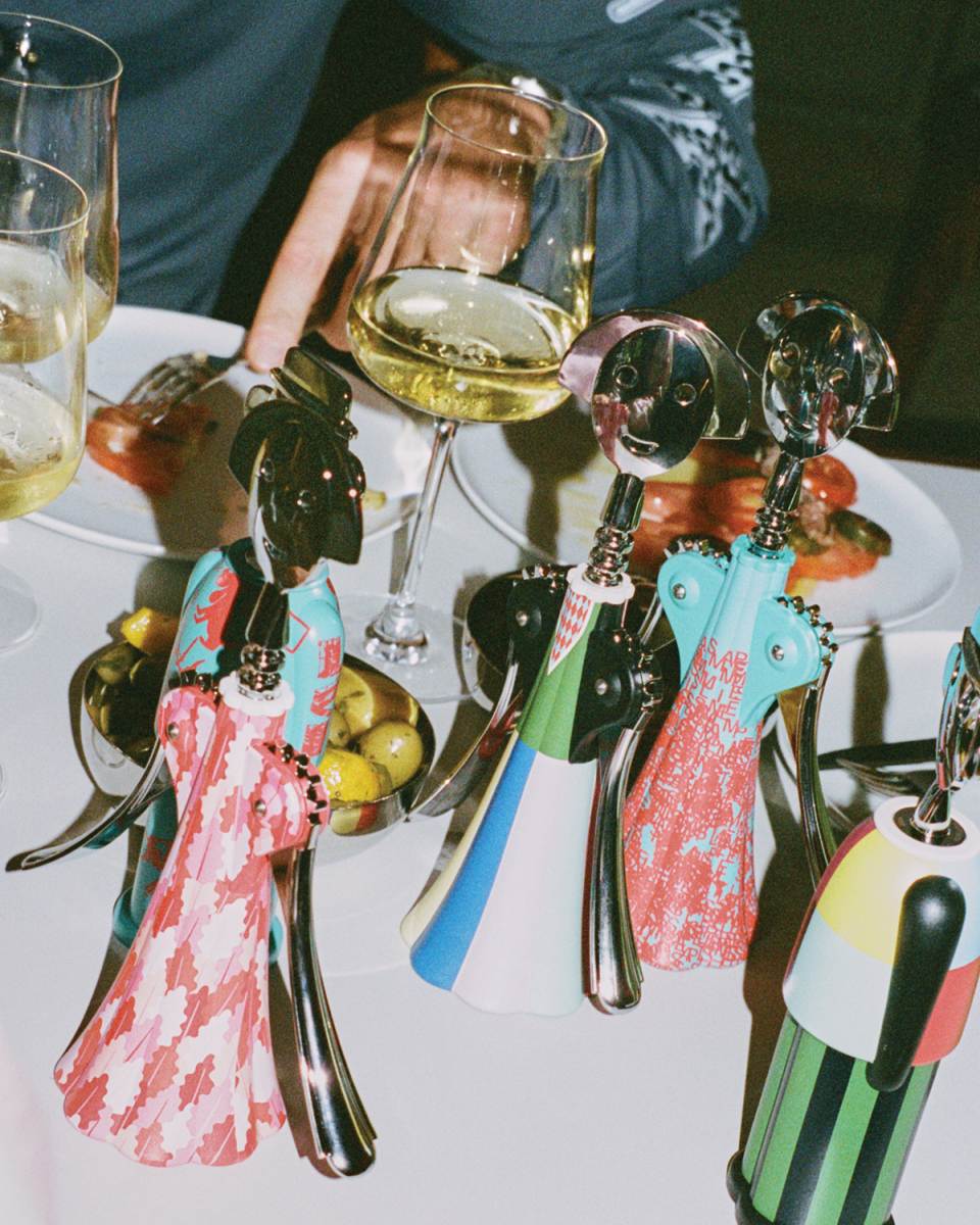 Mehrere bunte Anna G. Korkenzieher von Alessi auf einem gedeckten Tisch mit Weingläsern.