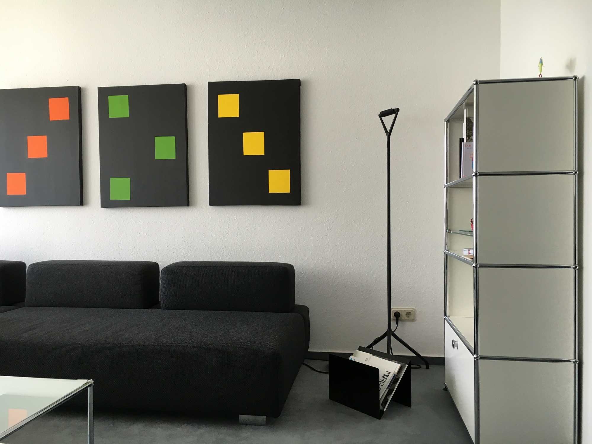 Schwarzer ZS01 M Zeitungsständer Stadie aus Metall neben einer Stehlampe und einem Sofa im Wohnzimmer.