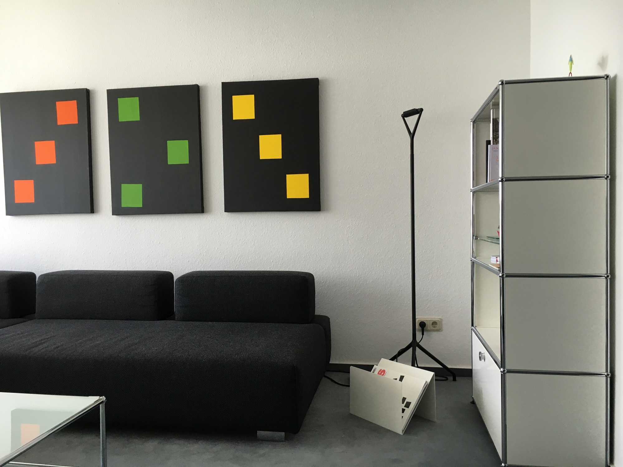 Moderner Wohnraum mit ZS01 M Zeitungsständer Stadie, schwarzem Sofa und minimalistischer Kunst an der Wand.