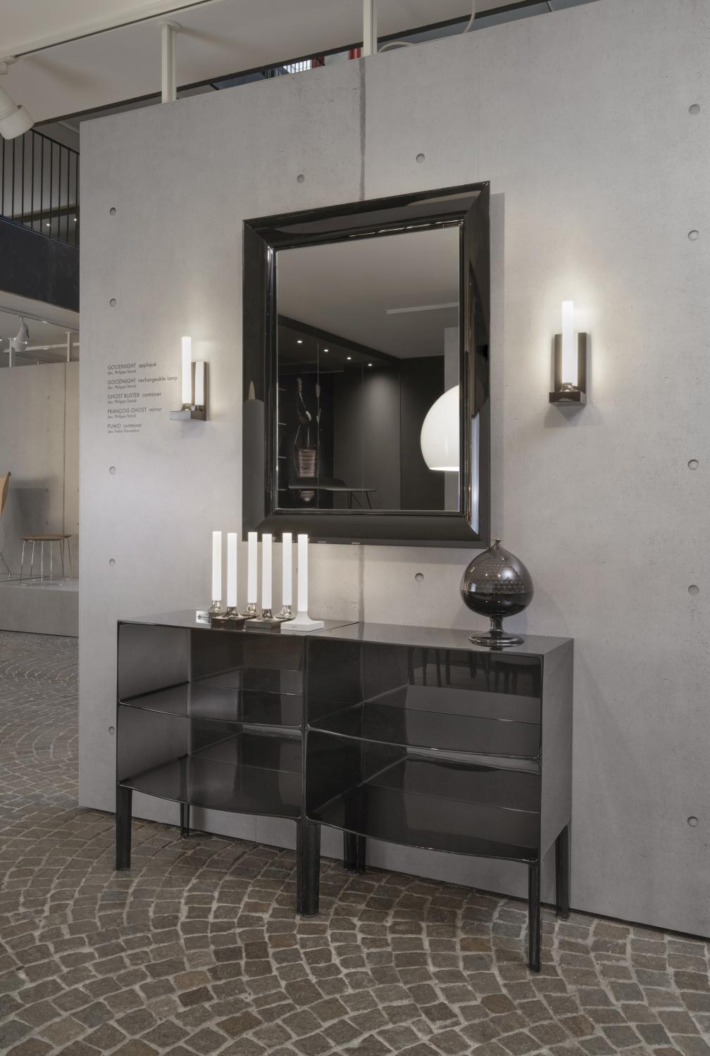 Goodnight Wandleuchte von Kartell, polierter Titan, an einer grauen Wand neben einem Spiegel.