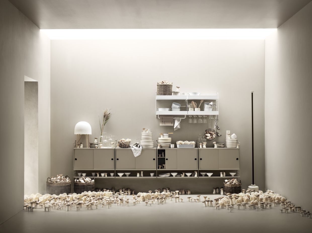 Beige String Furniture Küche mit Wandregal-System, Geschirr und Dekoration in einem hellen Raum.