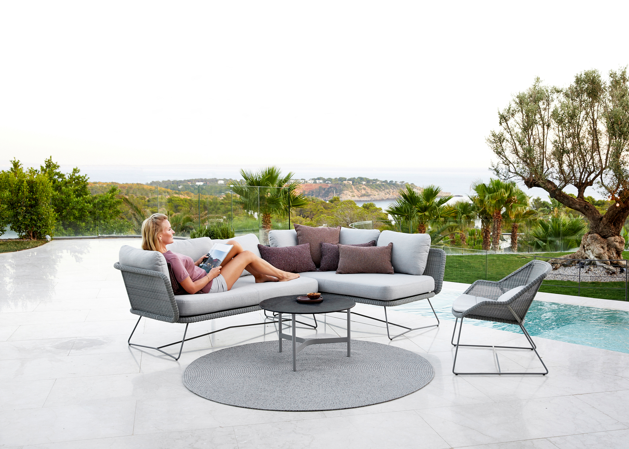 Frau entspannt auf Cane-Line Horizon Outdoor Sofa. Modulares Lounge-Möbel mit Blick auf Pool und Meer.
