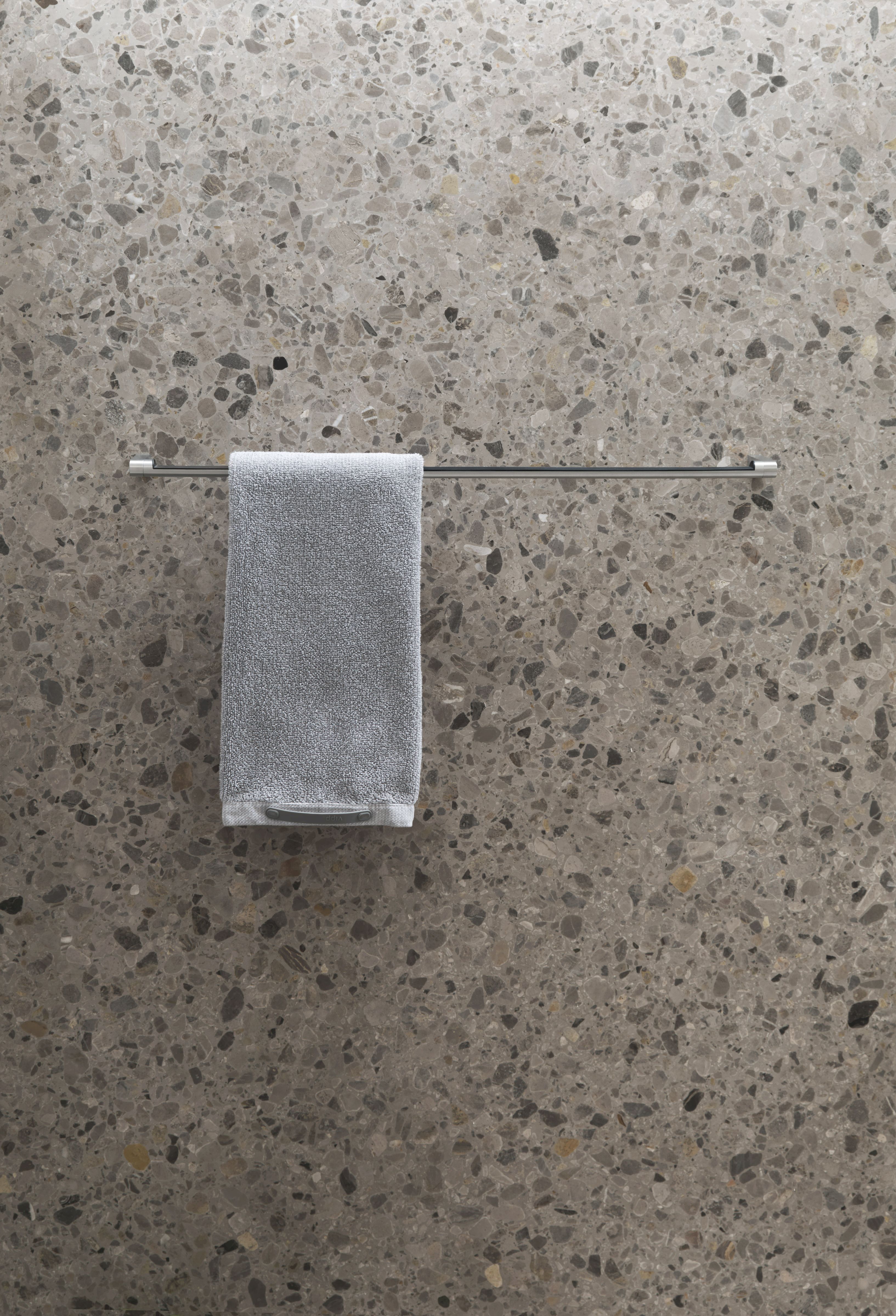 Silberne Vipp Handtuchstange mit grauem Handtuch vor einer grauen Wand mit Steinmuster.