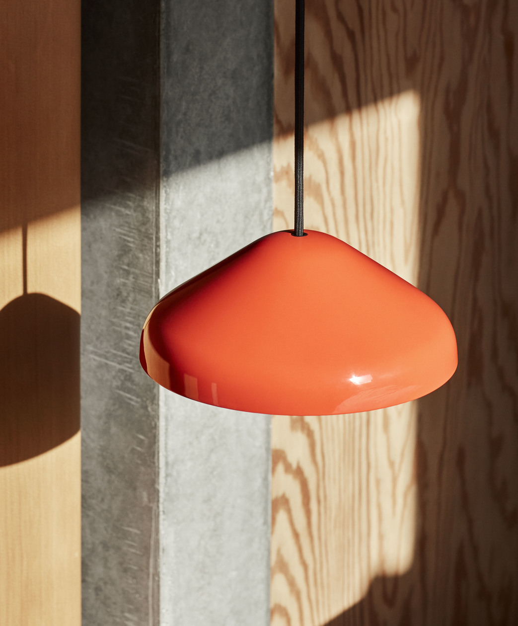 Orangefarbene Pao Steel Pendelleuchte von Hay mit schwarzem Kabel vor Holzwand im Sonnenlicht.
