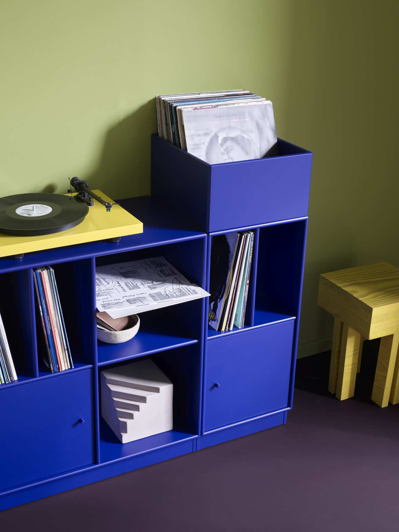Blaues Spin III Schallplattenregal mit Schallplatten, Plattenspieler und gelbem Hocker für moderne Vinyl-Aufbewahrung.