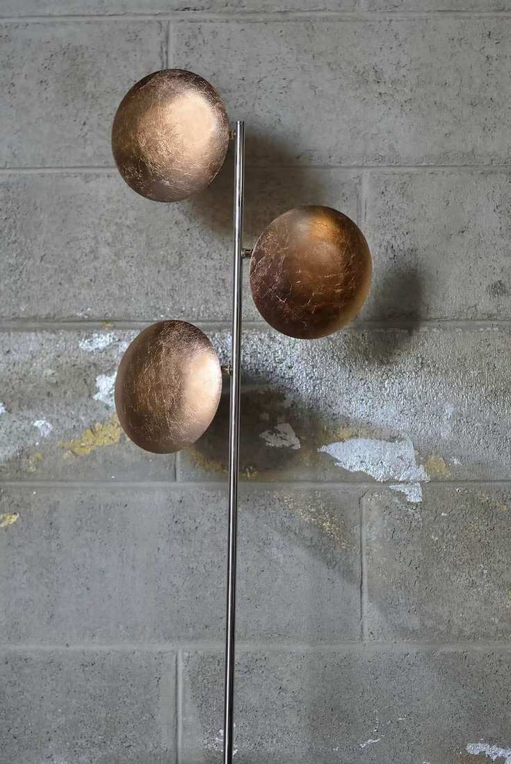 Lederam Stehleuchte mit drei goldenen Kugeln vor einer grauen Ziegelwand.