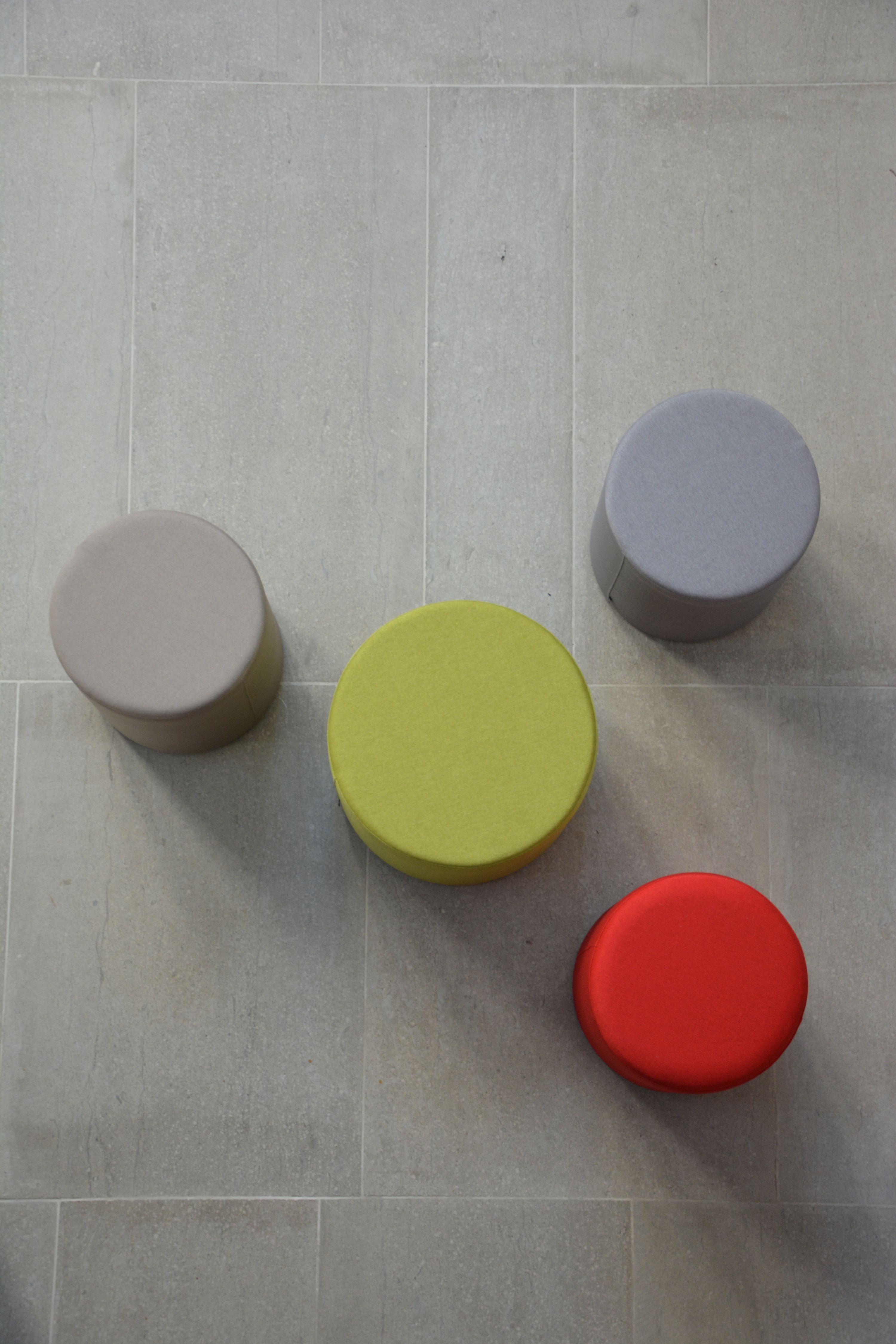 Draufsicht: Vier runde Poufs in Beige, Grün, Grau und Rot auf grauem Fliesenboden.