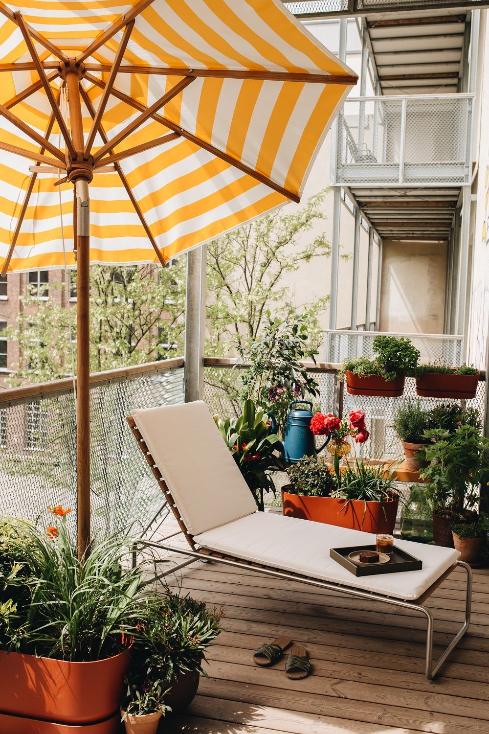 Weishäupl Teakschirm mit gelb-weiß gestreiftem Bezug spendet Schatten auf einem begrünten Balkon mit Liege.