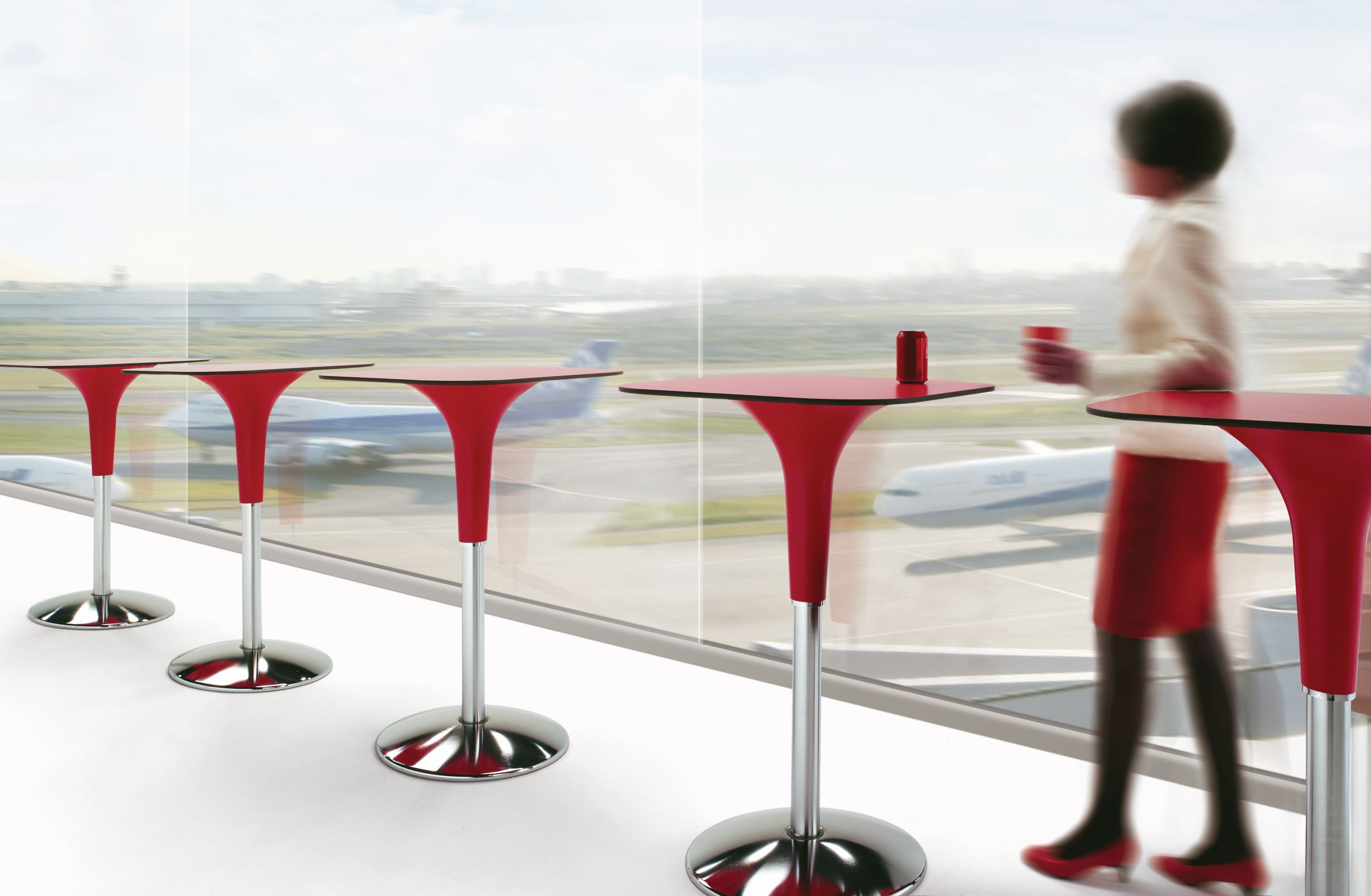 Zanziplano Tisch von Rexite, roter Stehtisch mit Frau und Getränk vor Fenster mit Flugzeugen.