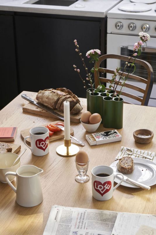 Frühstückstisch mit Brot, Eiern, Kaffeetassen mit Herzmotiv, grüner Vase und Kerze für gemütliche Morgenstunden.