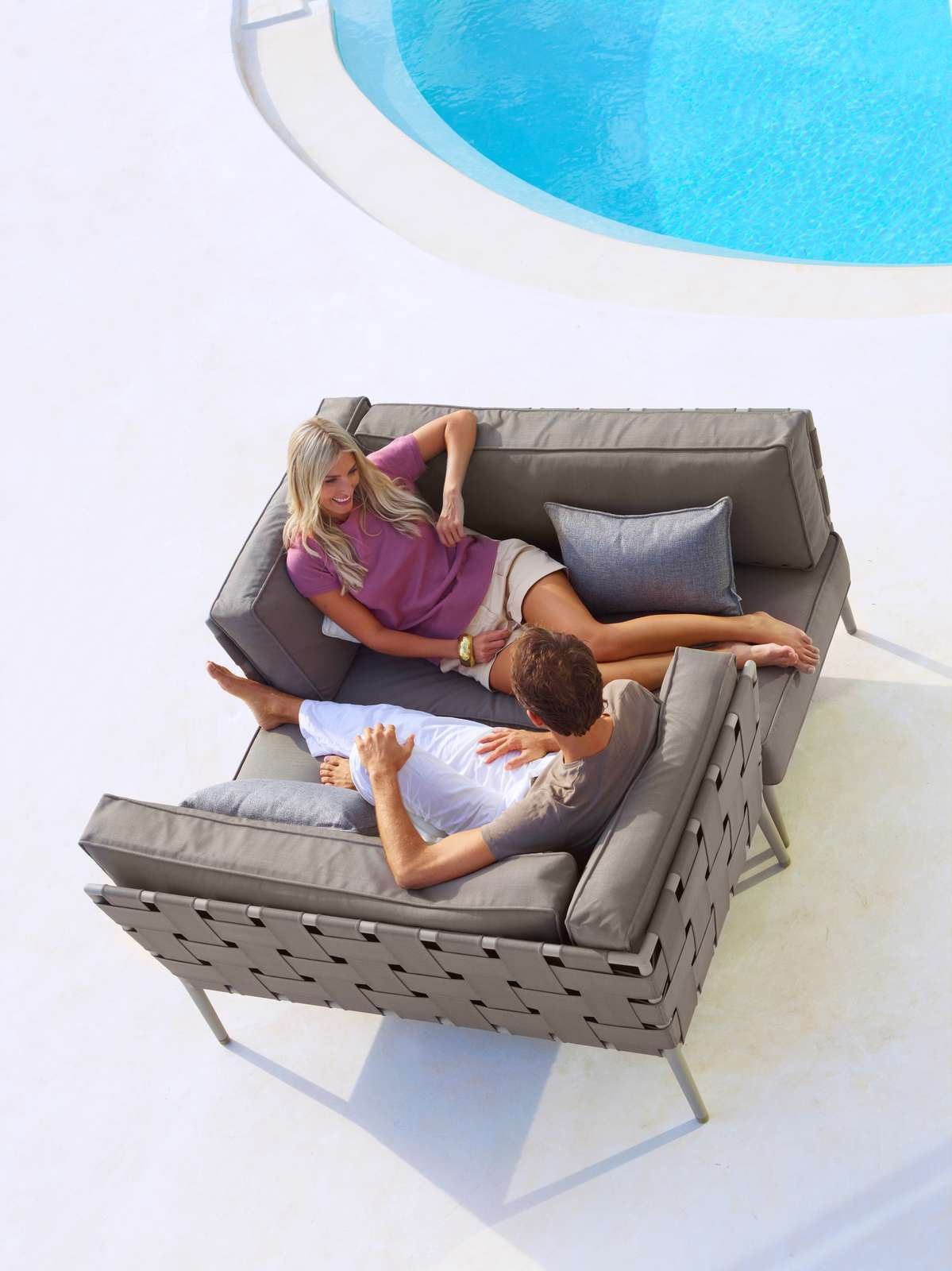 Paar entspannt auf hellgrauem Cane-Line Conic Modulsofa im Freien am Pool. Gartenmöbel für Terrasse.
