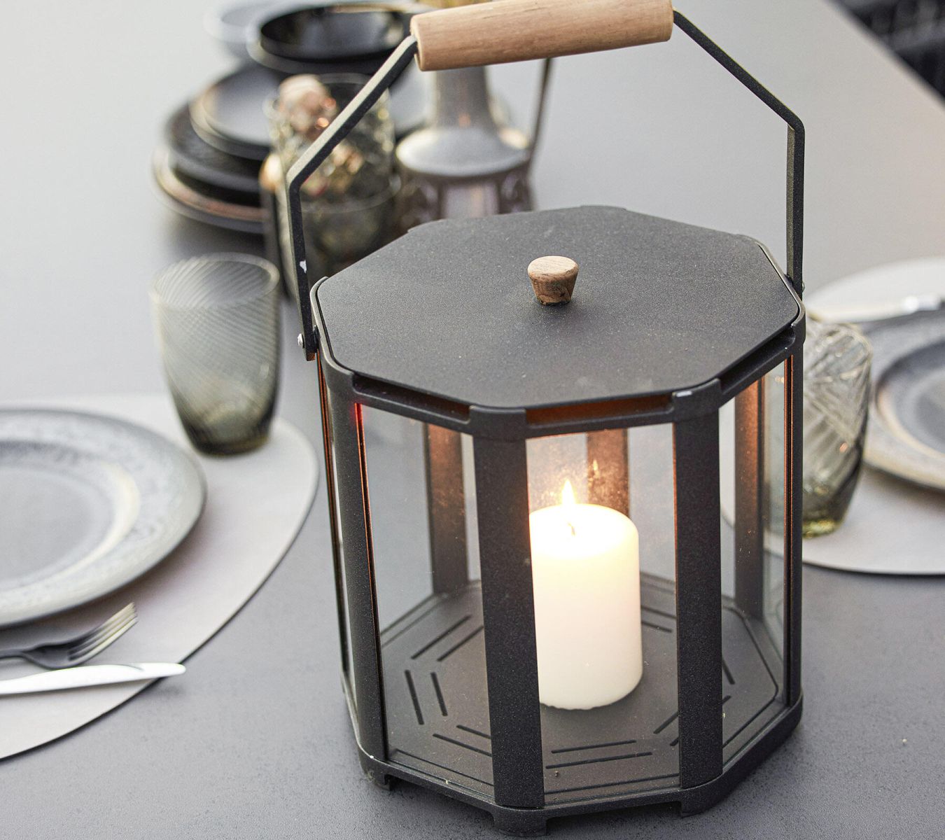 Lightlux Laterne Mini: Schwarze Outdoor-Laterne mit Teakholzgriff und brennender Kerze auf gedecktem Tisch.