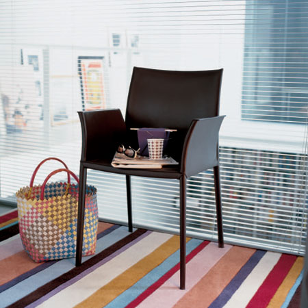 Brauner Zanotta Stuhl Lea ohne Armlehne auf buntem Teppich vor Fenster mit Jalousien. Designstuhl.