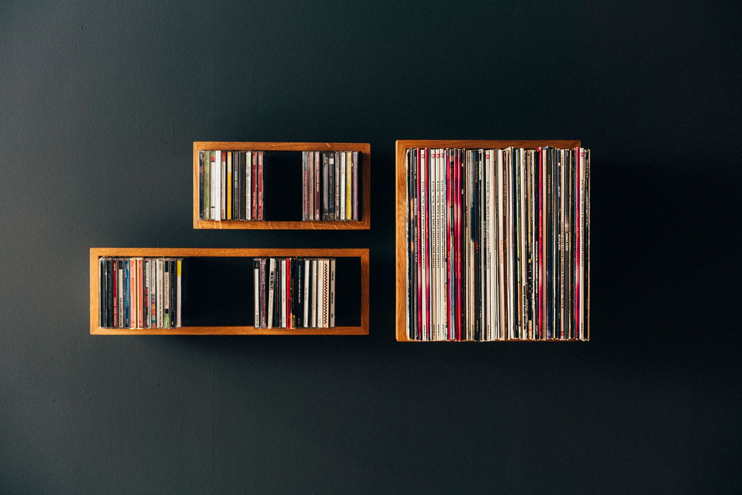 Drei hölzerne Schallplattenregale "das kleine b" an einer dunklen Wand, gefüllt mit Vinyl und CDs.