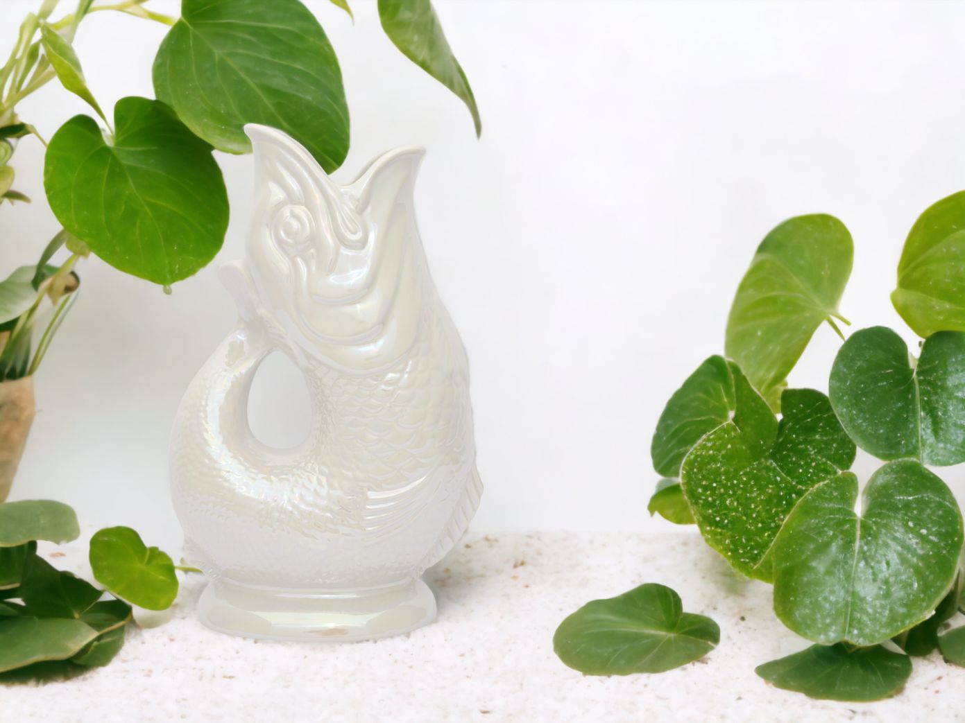 Gluckigluck Karaffe in Perlmutt-Optik, Fisch-Design, umgeben von grünen Pflanzen. Dekorative Wasserkaraffe für Tischdekoration.