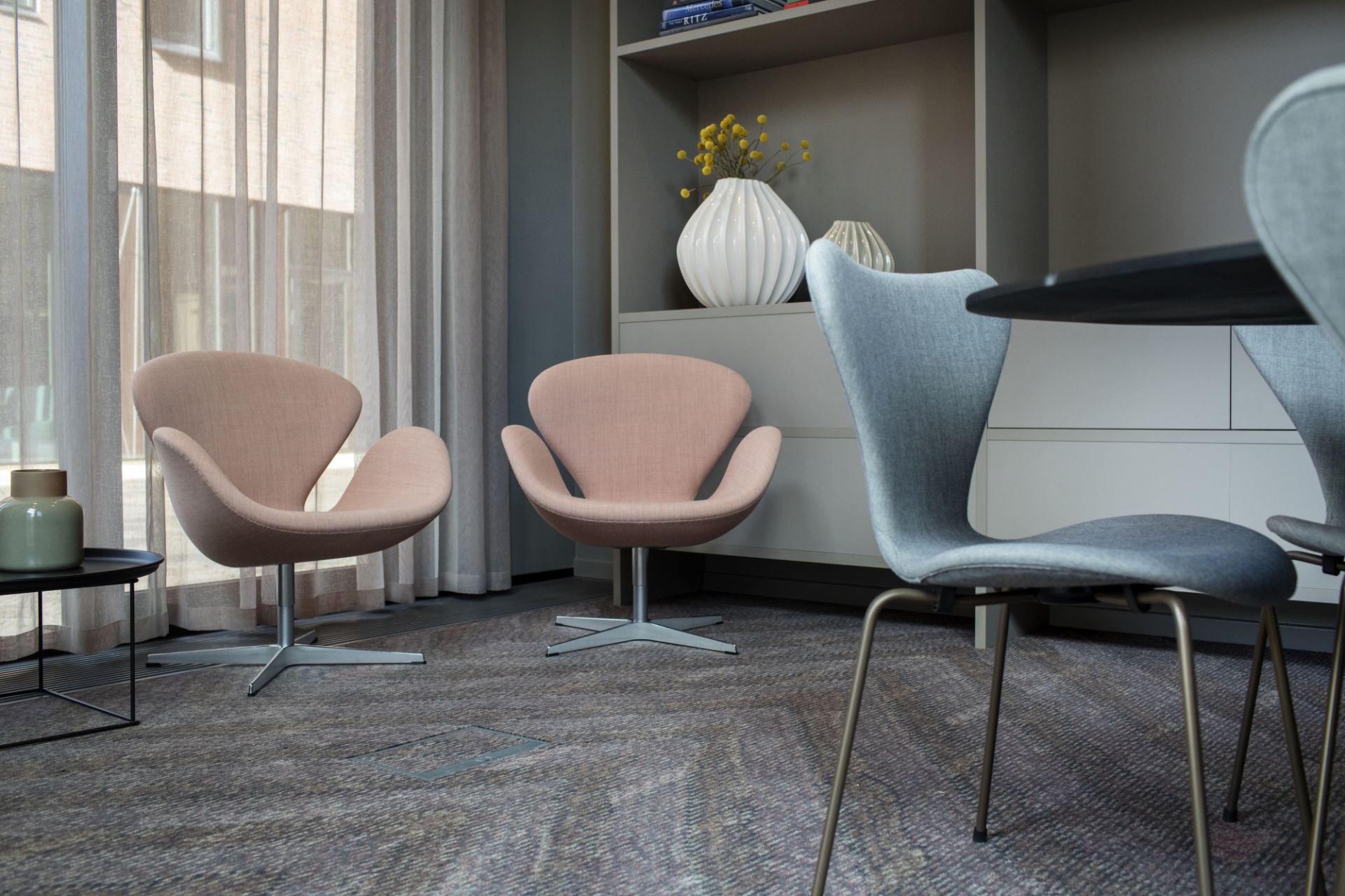 Zwei rosa Sessel und graue Esszimmerstühle in modernem Büro mit Regalen und Vorhängen.