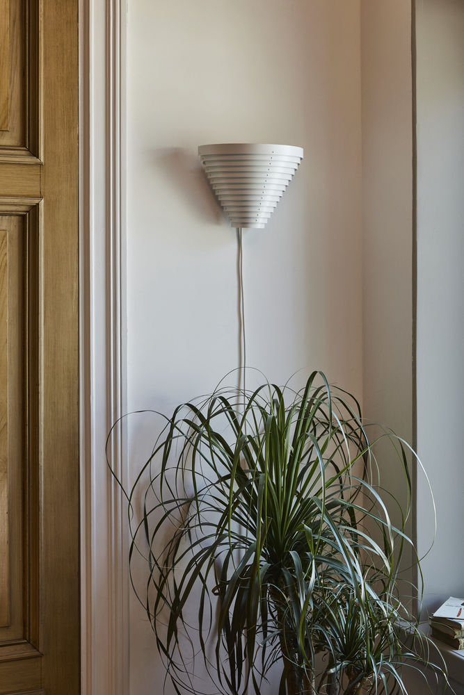 Weiße Artek A910 Wandleuchte mit konischem Schirm an einer Wand, neben einer Zimmerpflanze.
