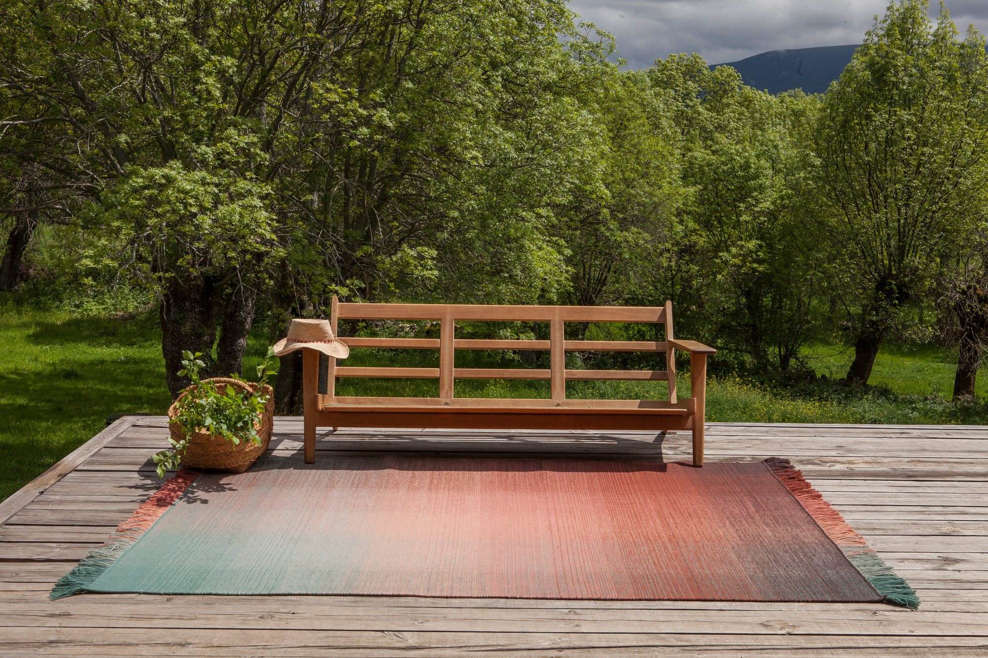 Nanimarquina Shade Teppich auf Holzterrasse mit Bank und Korb, Farbverlauf von Grün zu Rot.
