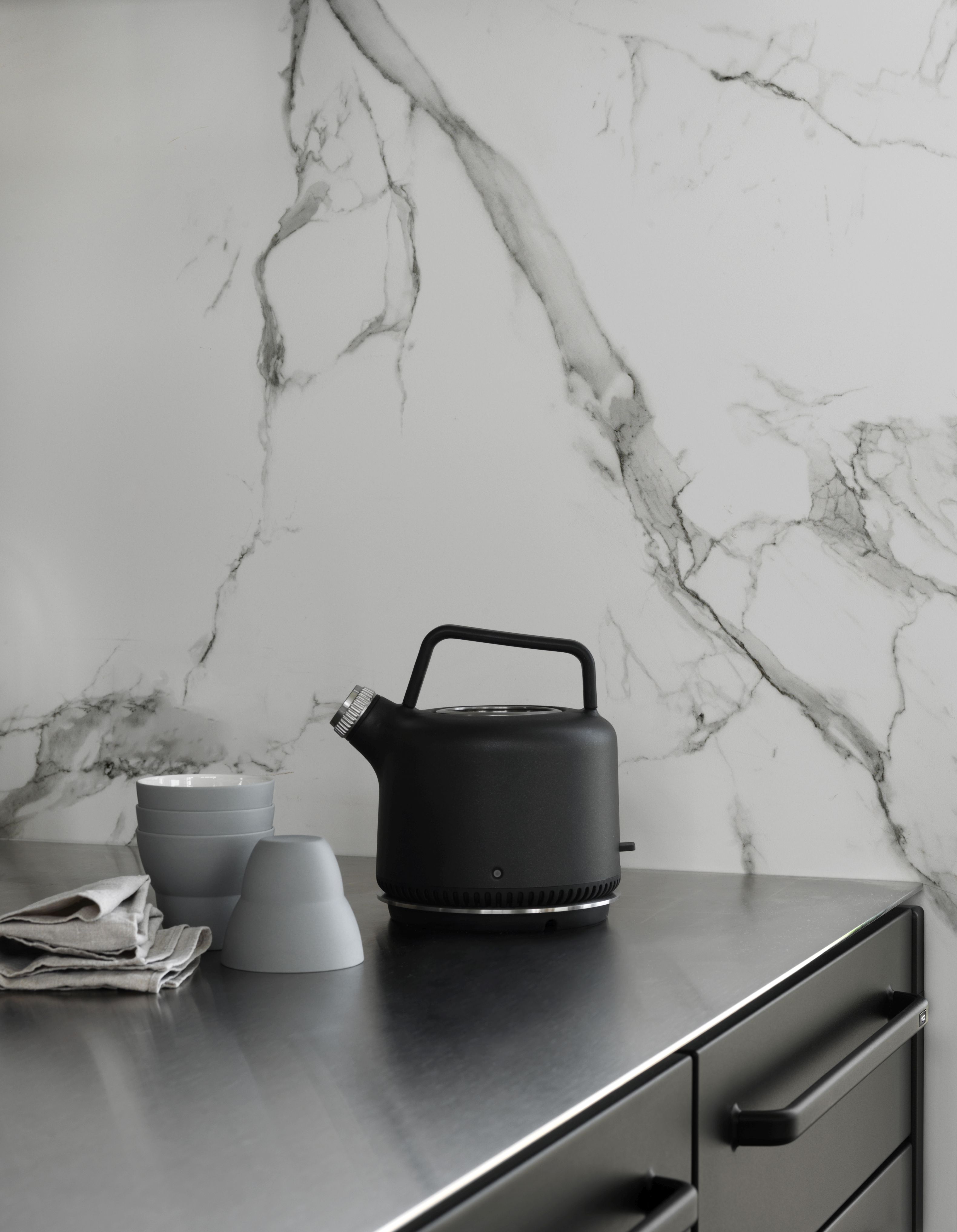Schwarzer Vipp Wasserkocher auf einer Küchentheke mit Geschirr vor einer Marmorwand.