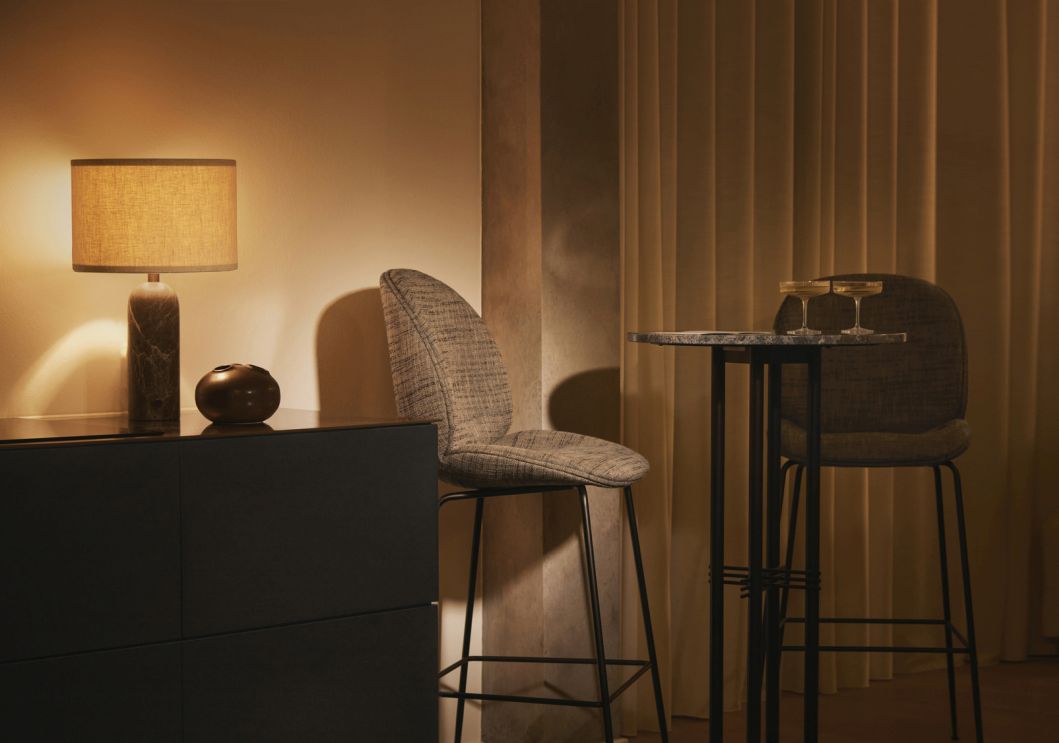 Gravity Tischleuchte mit weißem Schirm und Marmorfuß neben Kommode und Barhockern.