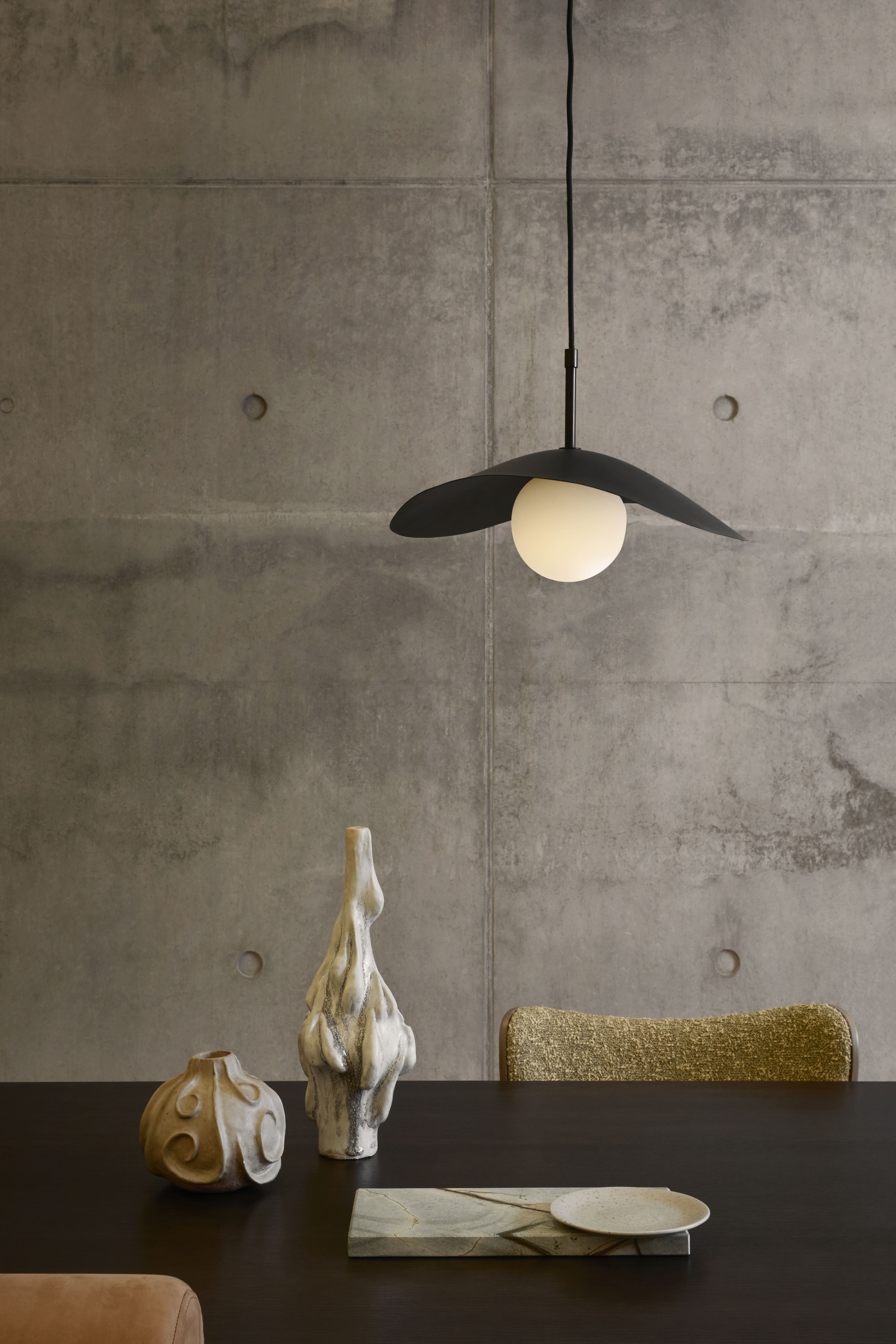 Fuji Pendelleuchte von Norr11 über Esstisch mit Deko-Objekten vor Betonwand. Design Lampe.