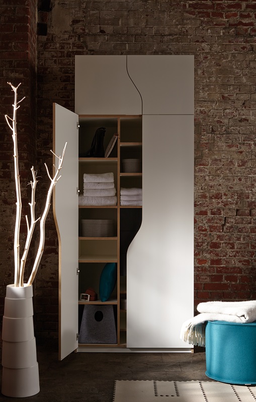 Weißer Müller Möbelwerkstätten Kleiderschrank mit offener Tür und Fächern vor einer Backsteinwand.