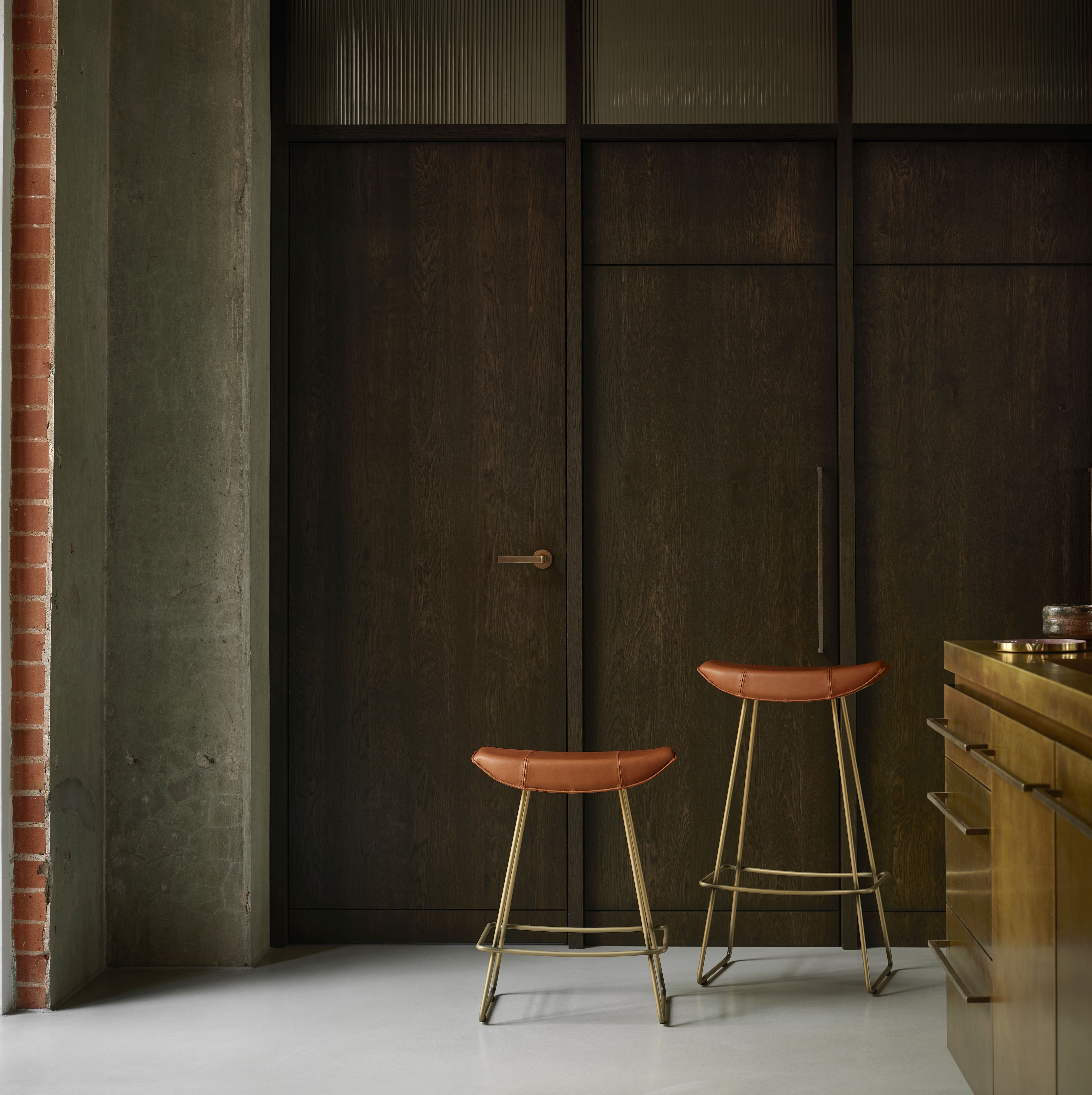 Zwei Kya Küchenhocker mit Drahtgestell und Lederbezug in Salbei vor dunkler Holzwand.