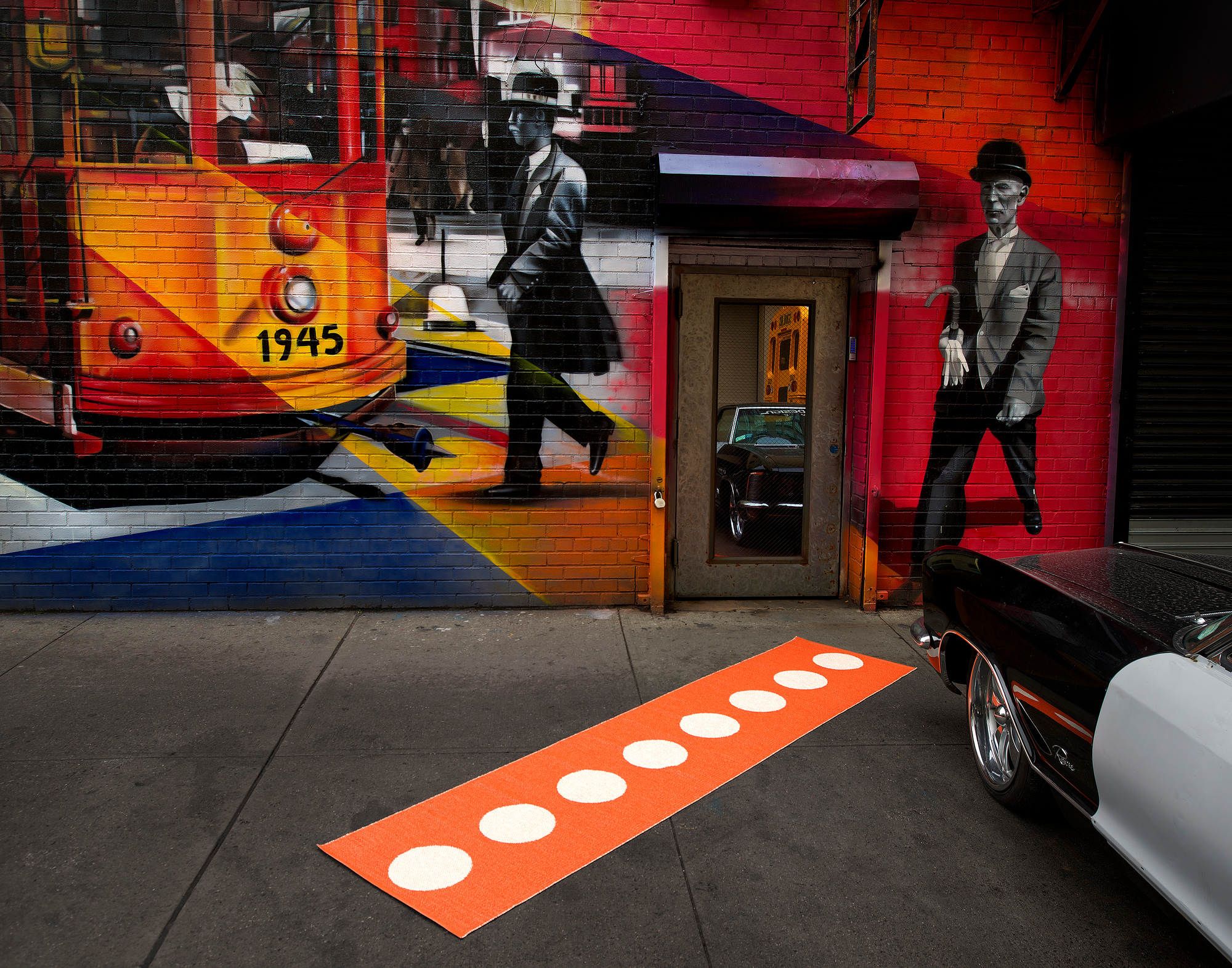 Vera Kunststoffteppich von Pappelina, orangefarbener Läufer mit weißen Punkten vor einer bemalten Hauswand in einer Großstadt.