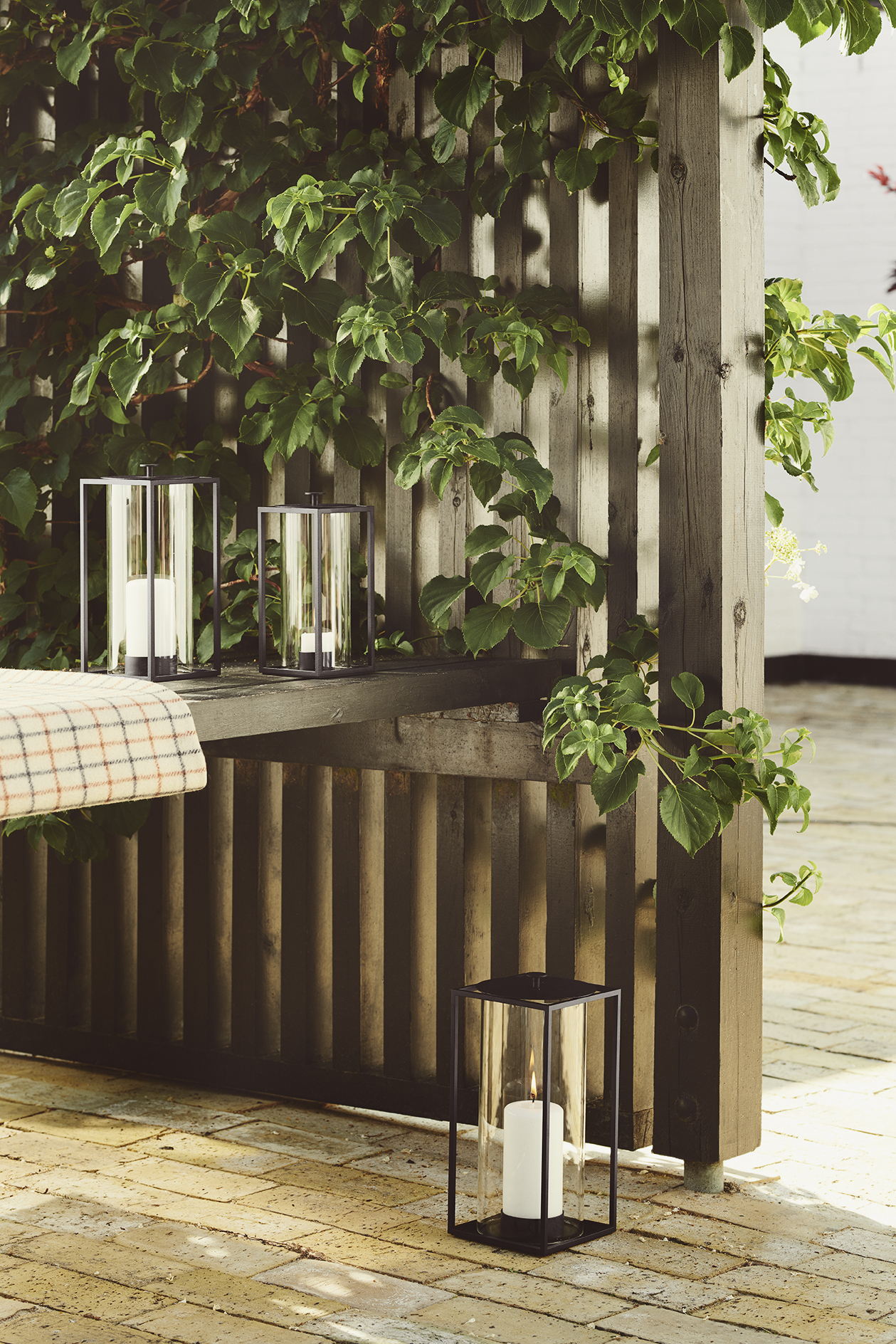 Drei schwarze Windlichter mit Kerzen auf einer Terrasse mit Holzwand und Pflanzen.