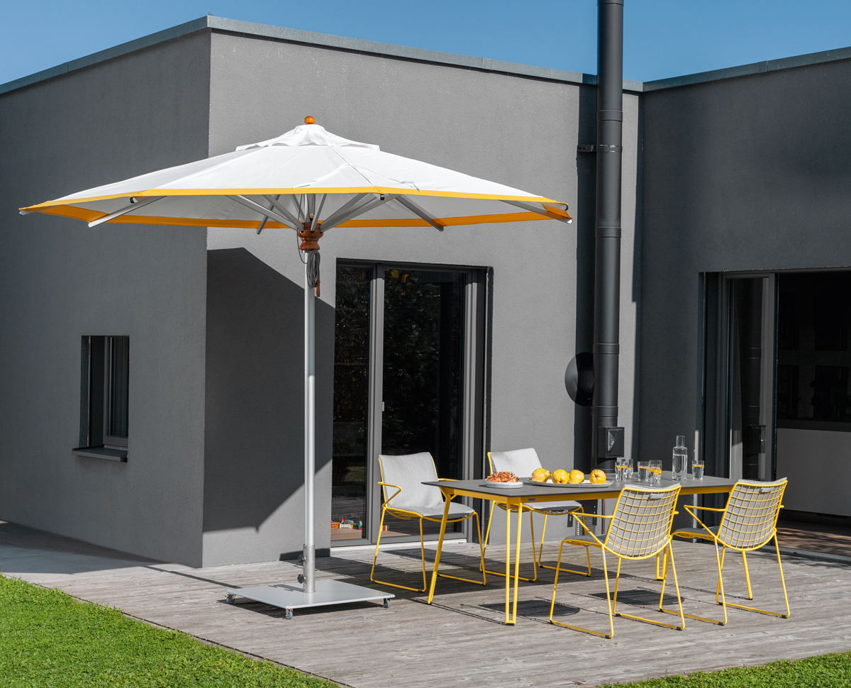Weißer Alusonnenschirm von Weishäupl mit gelbem Rand spendet Schatten auf einer Terrasse mit Gartenmöbeln.