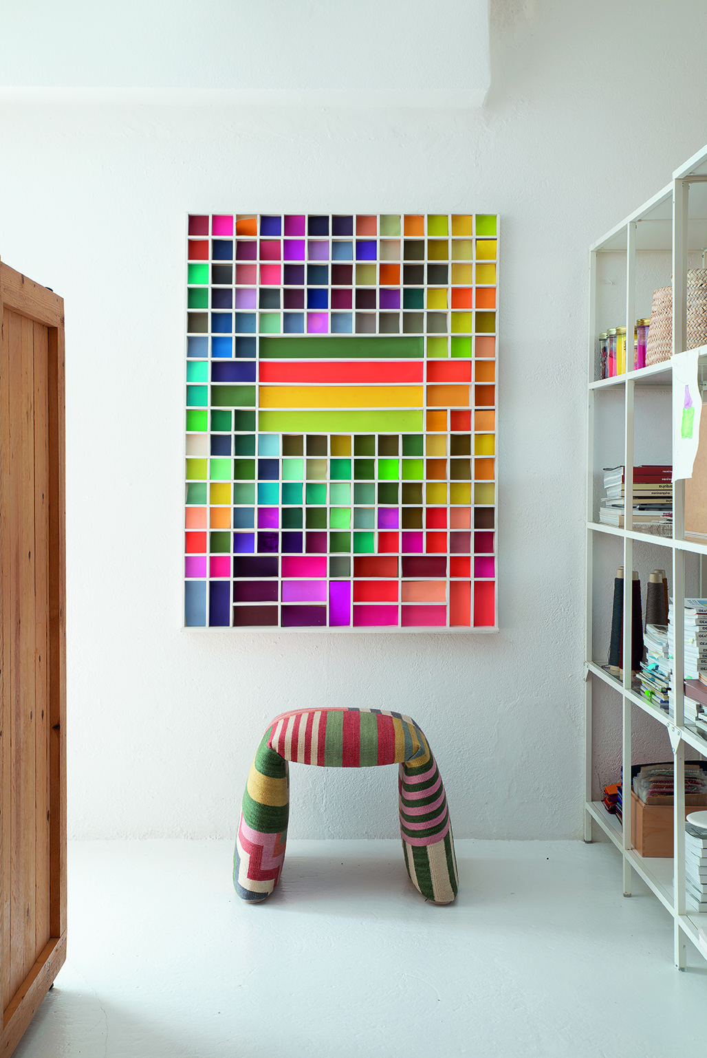 Nanimarquina Folded Pattern Bank: Gepolsterte Sitzbank mit bunten Streifen vor farbenfrohem Wandbild.