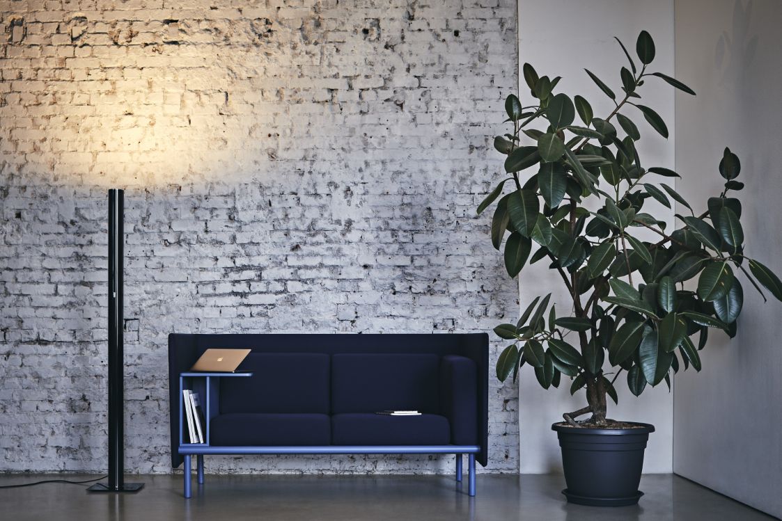 Floater Sofa von Cor, blaues 2-Sitzer Sofa mit Ablage, Stehlampe und Zimmerpflanze vor Backsteinwand.