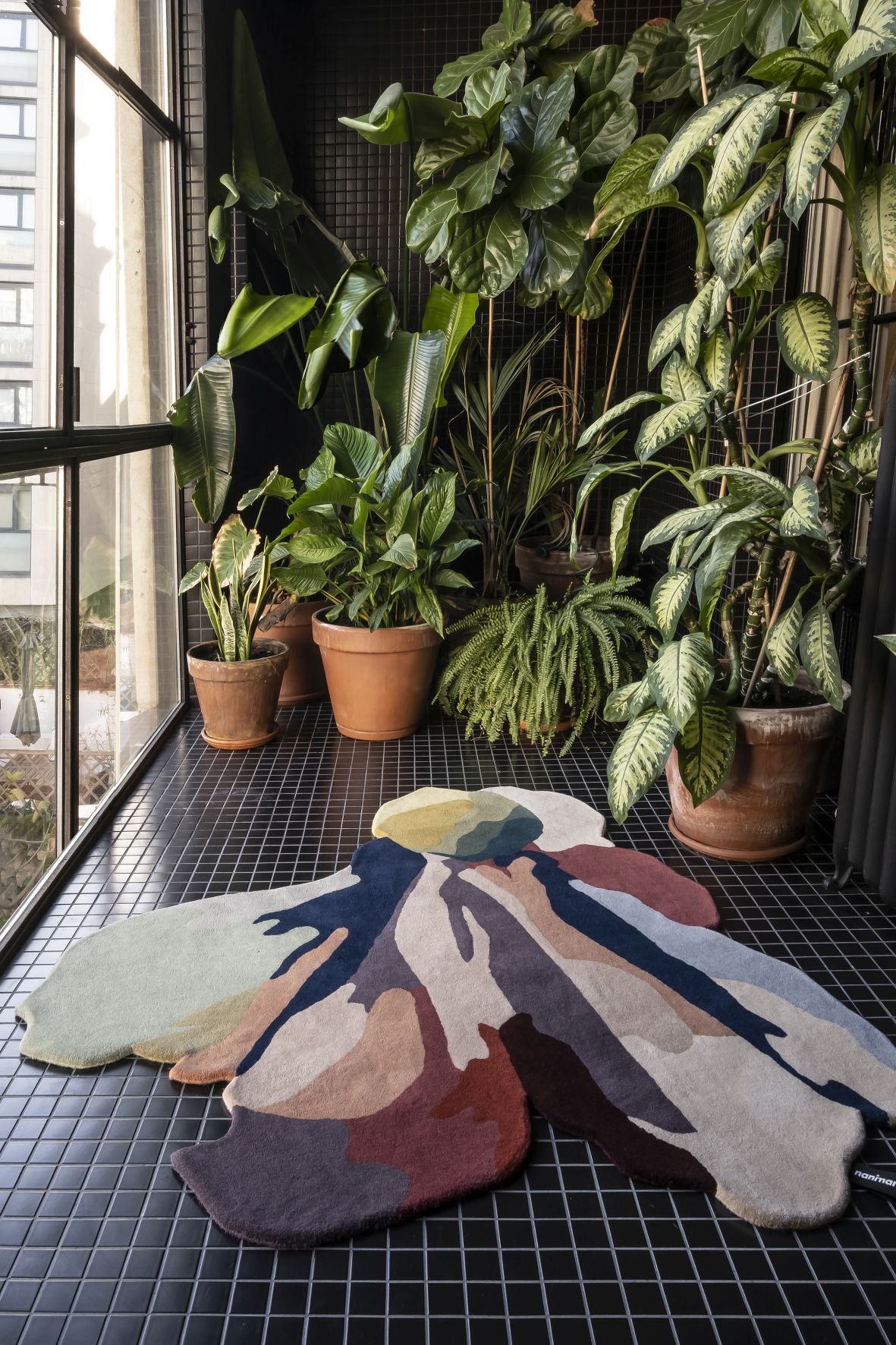 Nanimarquina Flora Bloom Teppich, 150x166 cm, abstrakte Blütenform in bunten Erdtönen auf dunklem Fliesenboden.