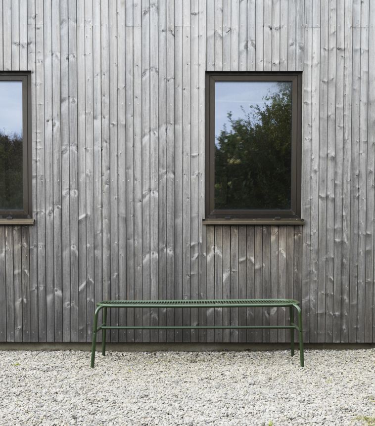 Grüne Vig Bench Bank von Normann Copenhagen für den Außenbereich vor grauer Holzwand.