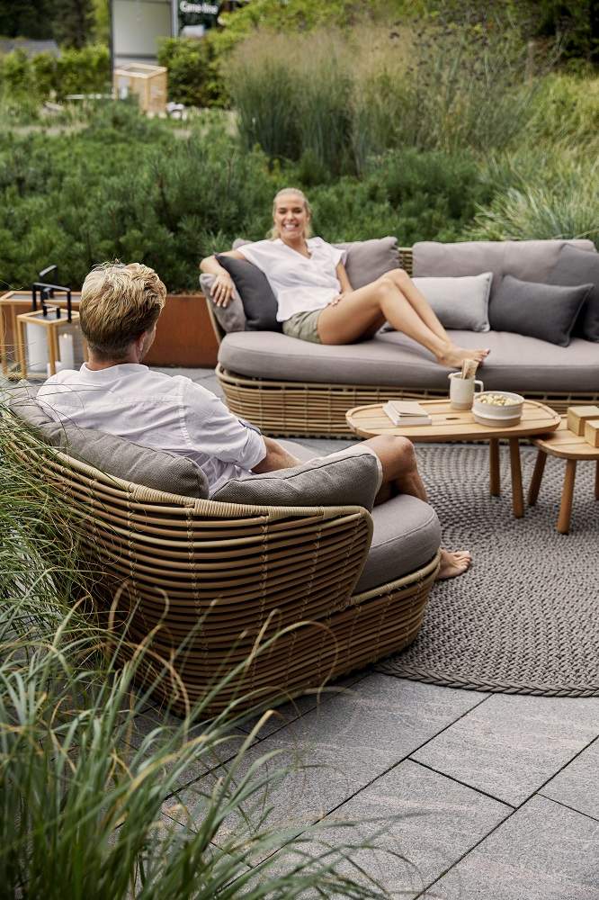 Basket Outdoor Sofa von Cane-Line: Zwei Personen entspannen auf stilvollen Rattan Gartenmöbeln im Freien.