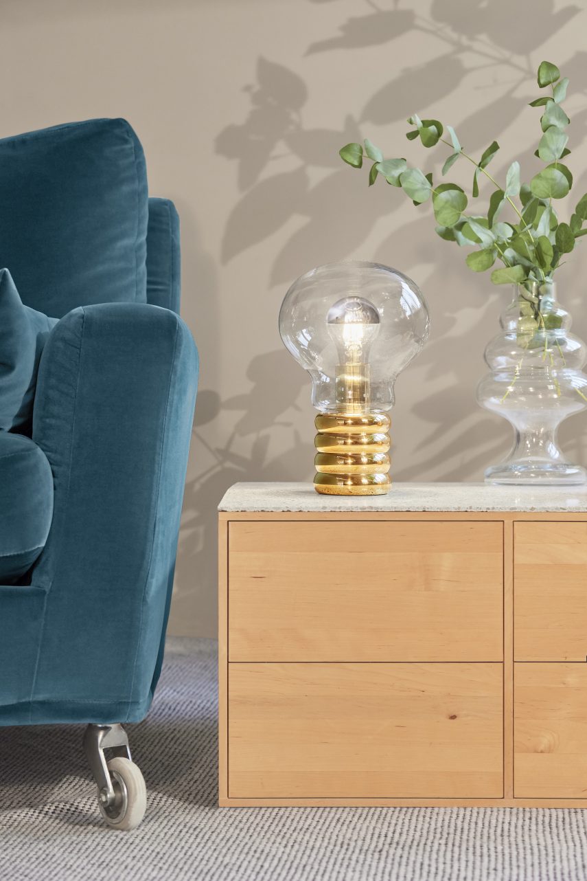 Ingo Maurer Bulb Tischleuchte aus Messing und Glas auf einem Nachttisch neben einem Sessel.