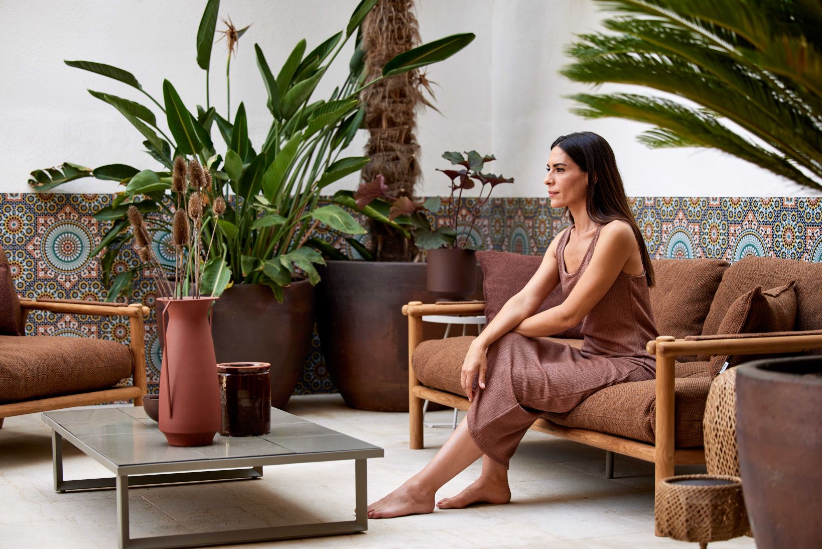 Frau entspannt auf Cane-Line Sticks Outdoor Sofa mit Kissen in einem begrünten Innenhof.