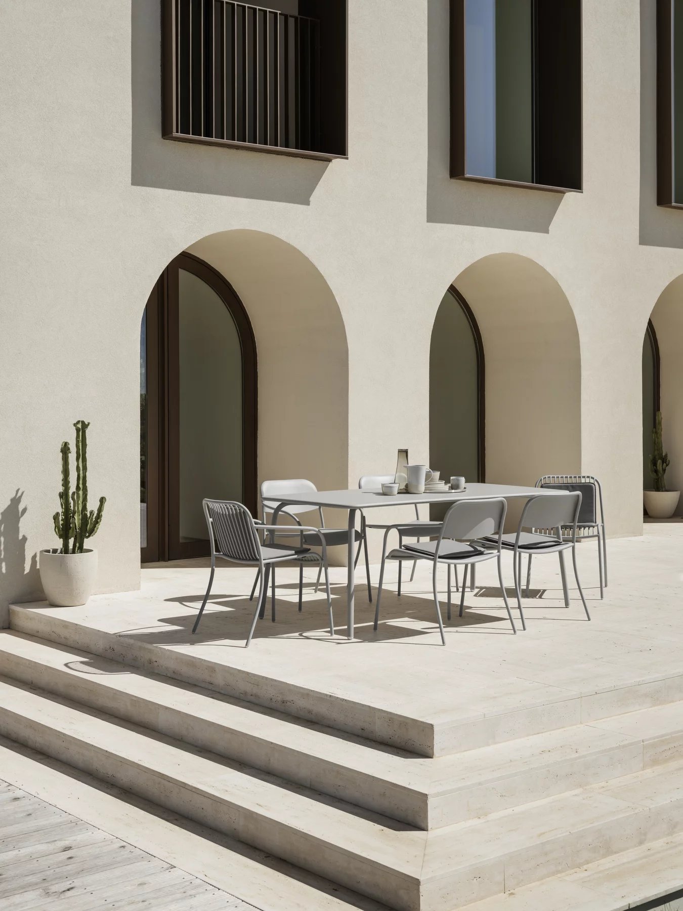 Blomus Yua Esstisch Outdoor mit Stühlen auf einer Terrasse vor einem Gebäude mit Bögen.