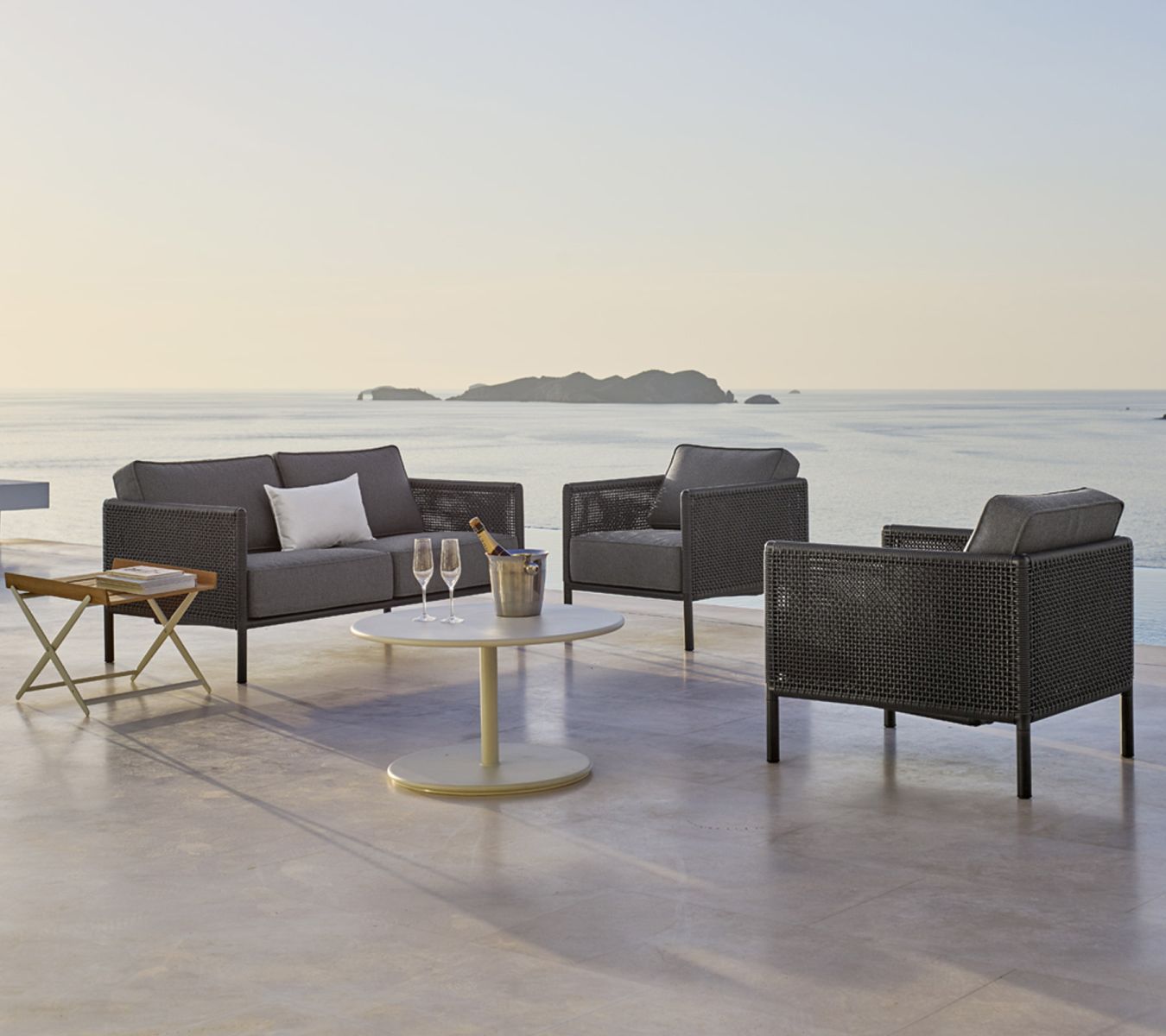 Encore Sofa von Cane-Line mit AirTouch Kissen, Gartensessel und Tisch auf Terrasse mit Meerblick.