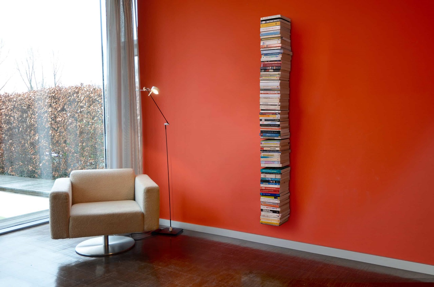Booksbaum Wandregal: Vertikales Bücherregal an oranger Wand, gefüllt mit Büchern, neben Sessel und Lampe.