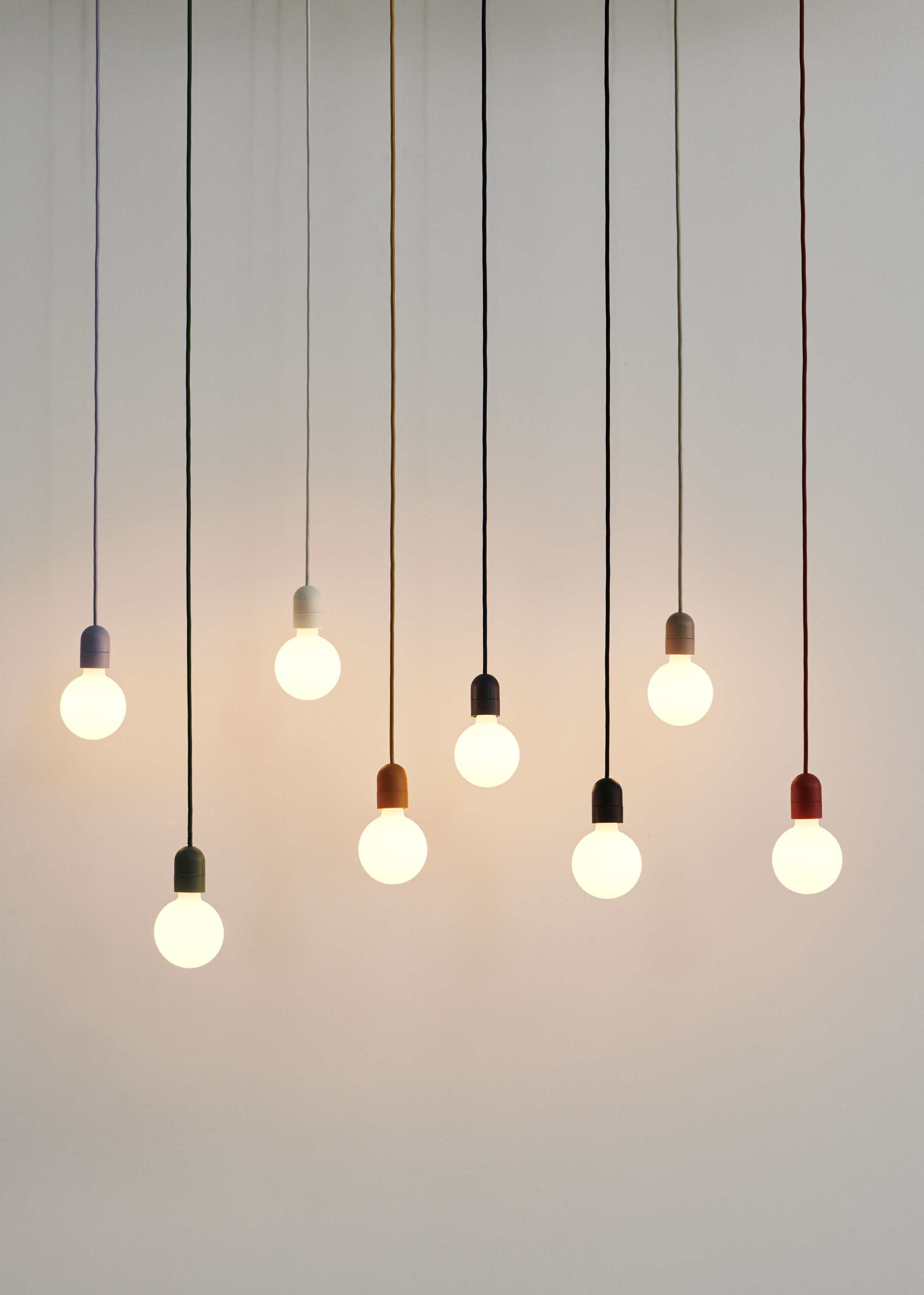 Mehrere Hay Pendelleuchten mit farbigen Textilkabeln und Glühbirnen vor hellem Hintergrund.