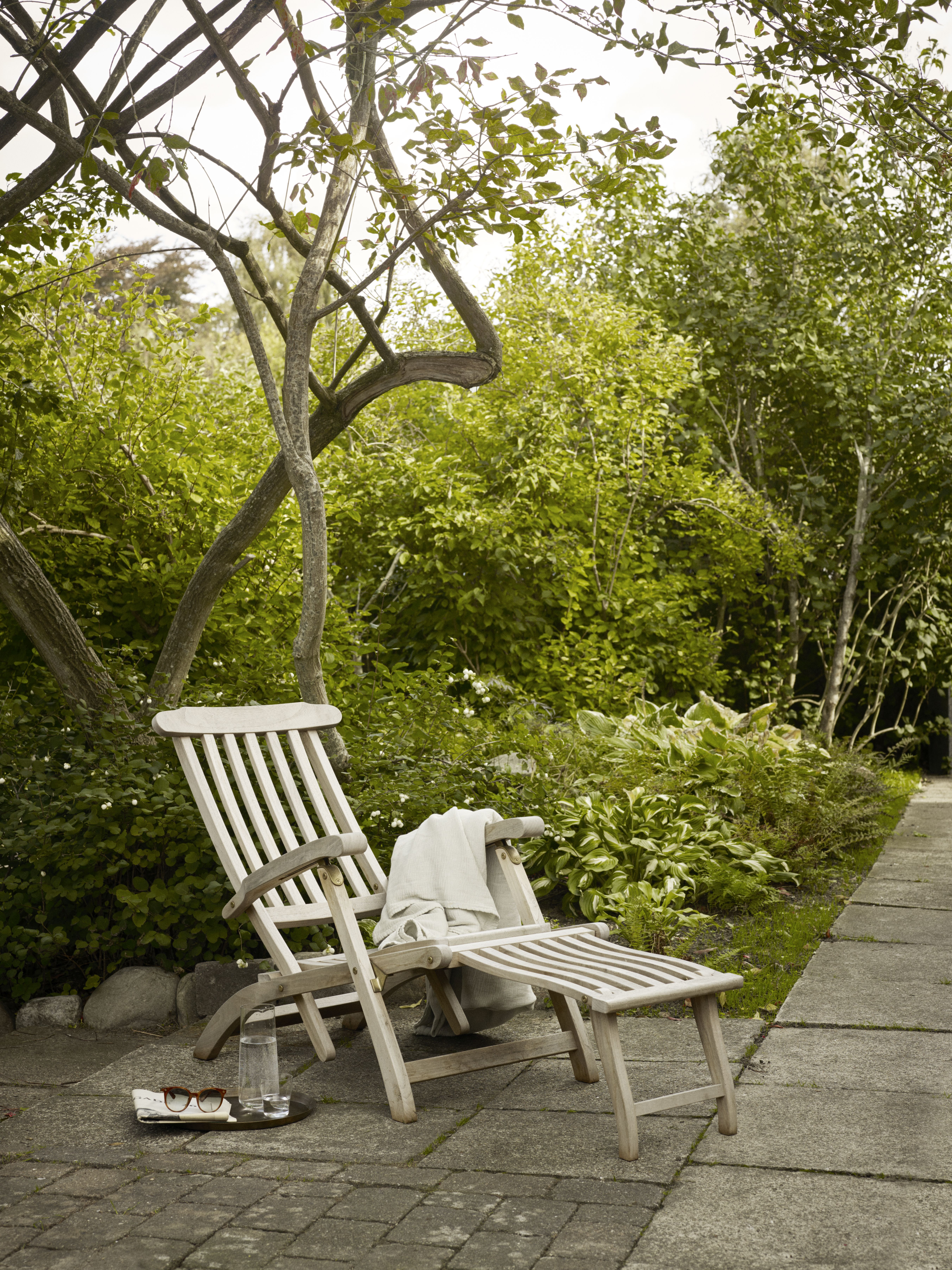 Holzliegestuhl "Steamer Deck Chair" im Garten, Outdoor-Möbel mit Fußteil, Handtuch, Getränk und Sonnenbrille.