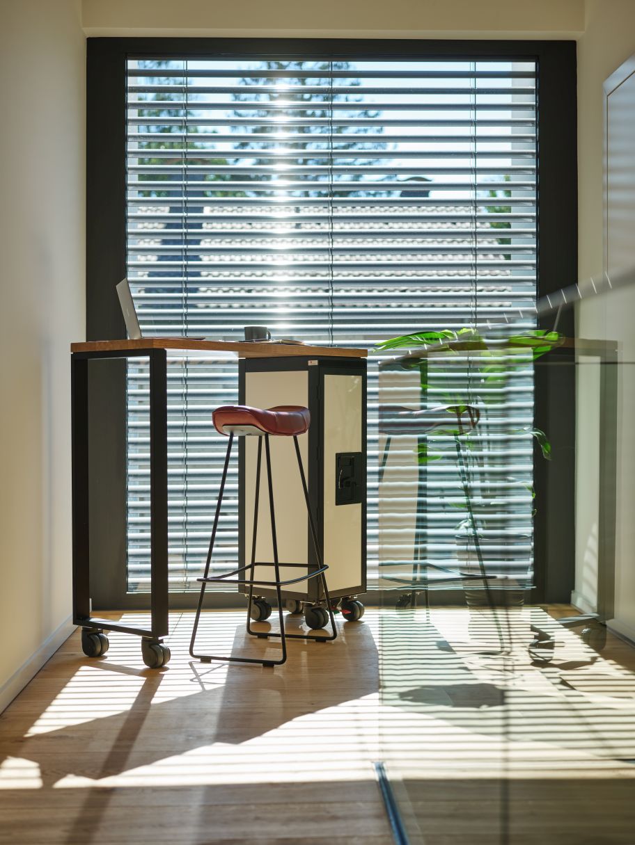 Schwarzer Janua Hightable Tisch mit Bordbar und rotem Barhocker vor Fenster mit Jalousien.