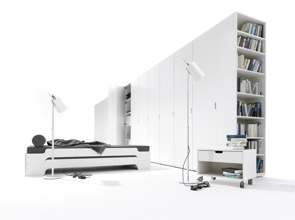 Weißer Müller Möbelwerkstätten Nachttisch mit Schublade und Rollen vor weißem Kleiderschrank und Bücherregal.