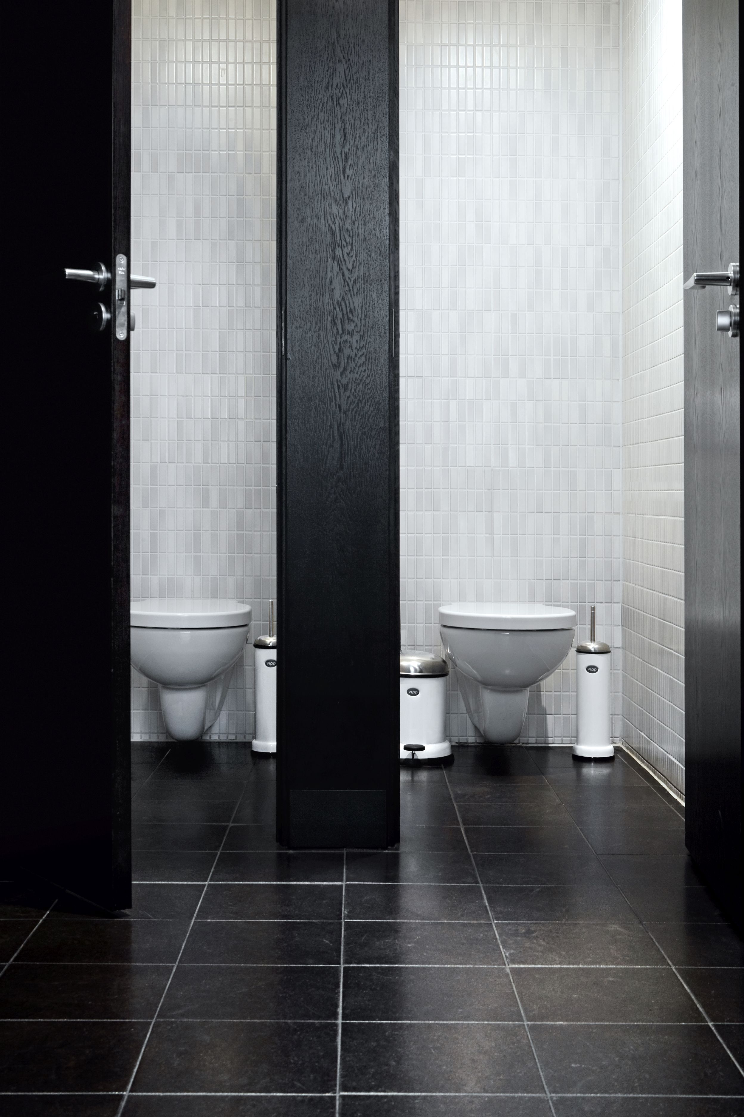 Zwei Toilettenräume mit weißen WCs, schwarzen Fliesen und jeweils einer weißen Vipp WC-Bürste.