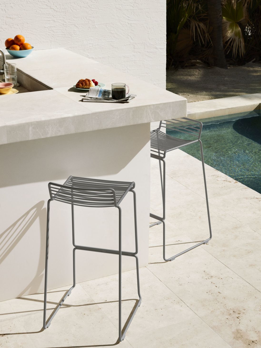 Zwei graue Hee Barhocker von Hay, feuerverzinkt, im Außenbereich neben einem Pool.